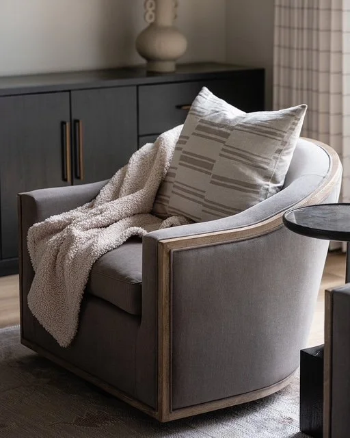 A cozy living room corner with a gray armchair, a beige textured blanket, and a decorative pillow. A black side table is beside the chair, and there is a dark cabinet and a white lamp in the background.