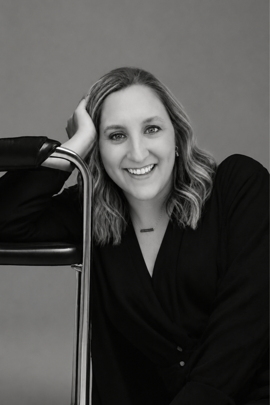 Black and white portrait of a smiling woman with wavy hair, wearing a dark top, resting her head on her hand, seated beside a metal chair with a cushion.