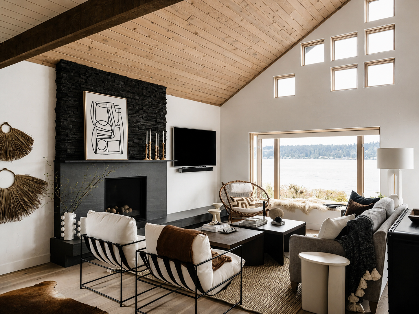 Living room with a vaulted ceiling, wooden planks, and multiple windows overlooking a body of water. Contains a black fireplace, wall art, a flat-screen TV, a mix of modern and rustic furniture including a sofa, chairs, and decorative items.