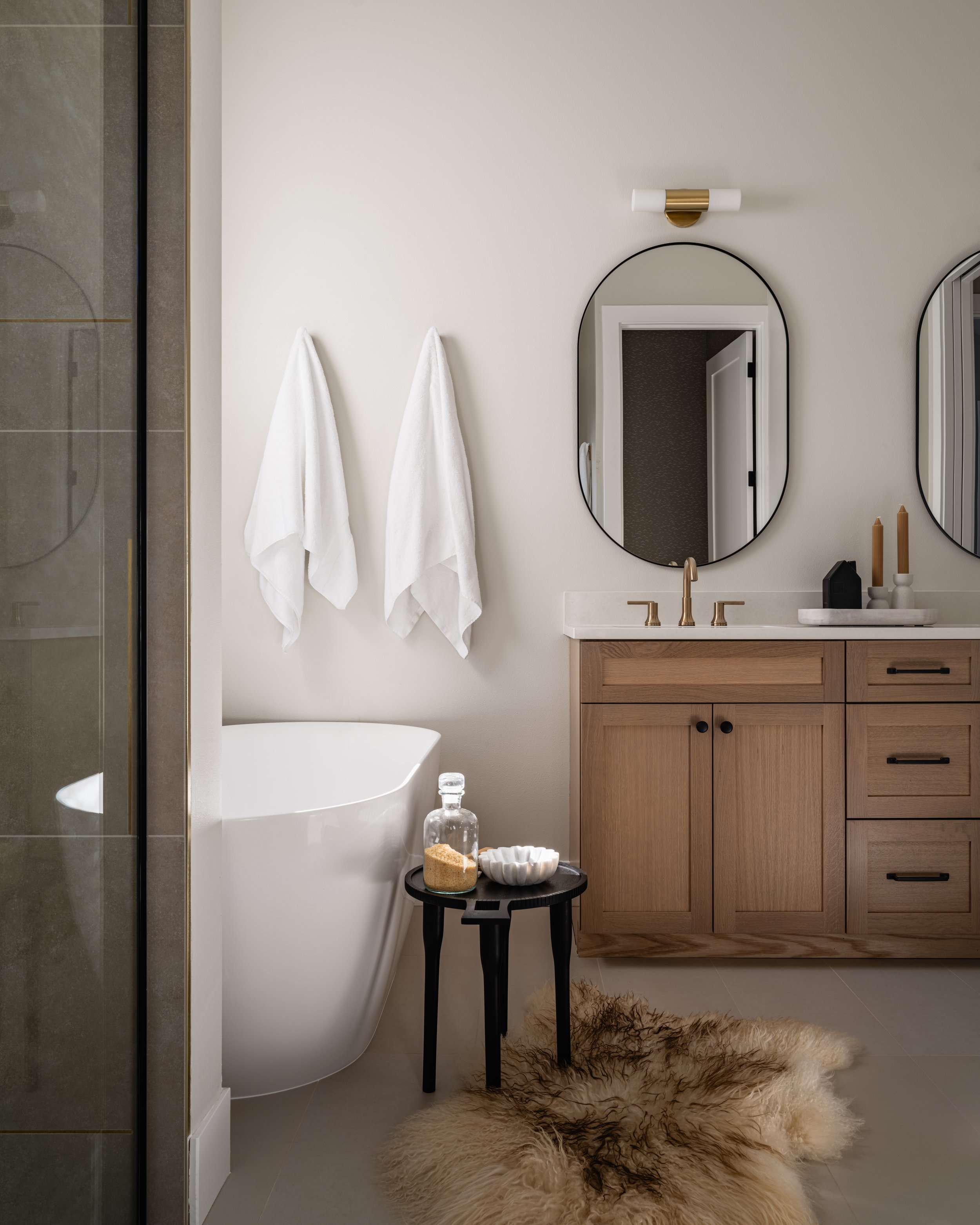 Modern bathroom with a wood vanity, oval mirrors, a black side table with a jar of bath salts and a bowl, white towels hanging on the wall, a textured beige rug on the floor, and part of a glass shower enclosure on the left.