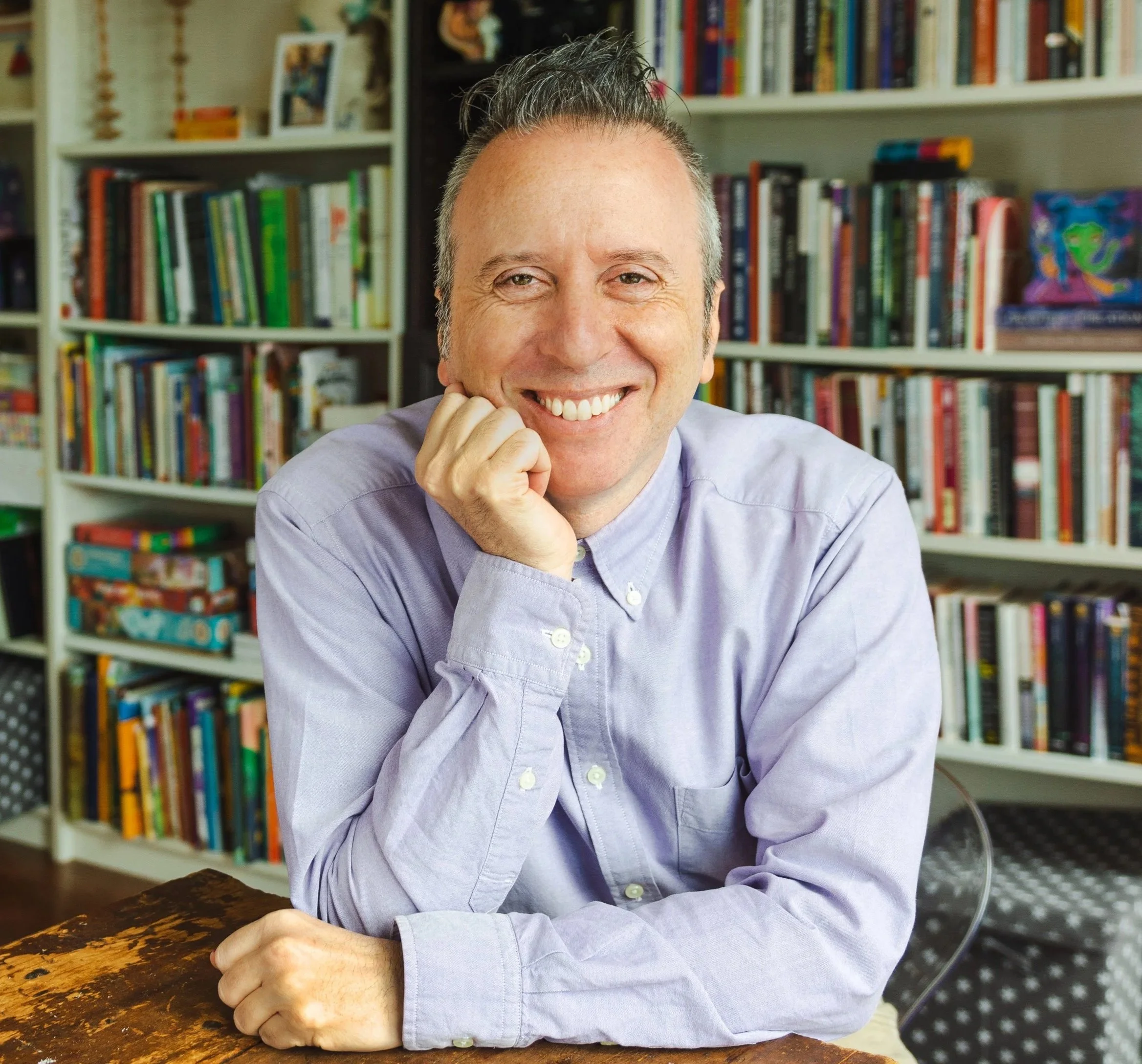 Cory Silverberg smiling, wearing a light purple button-up shirt, resting his chin on his hand and sitting at a wooden table in a room with white shelves filled with colorful books in the background.