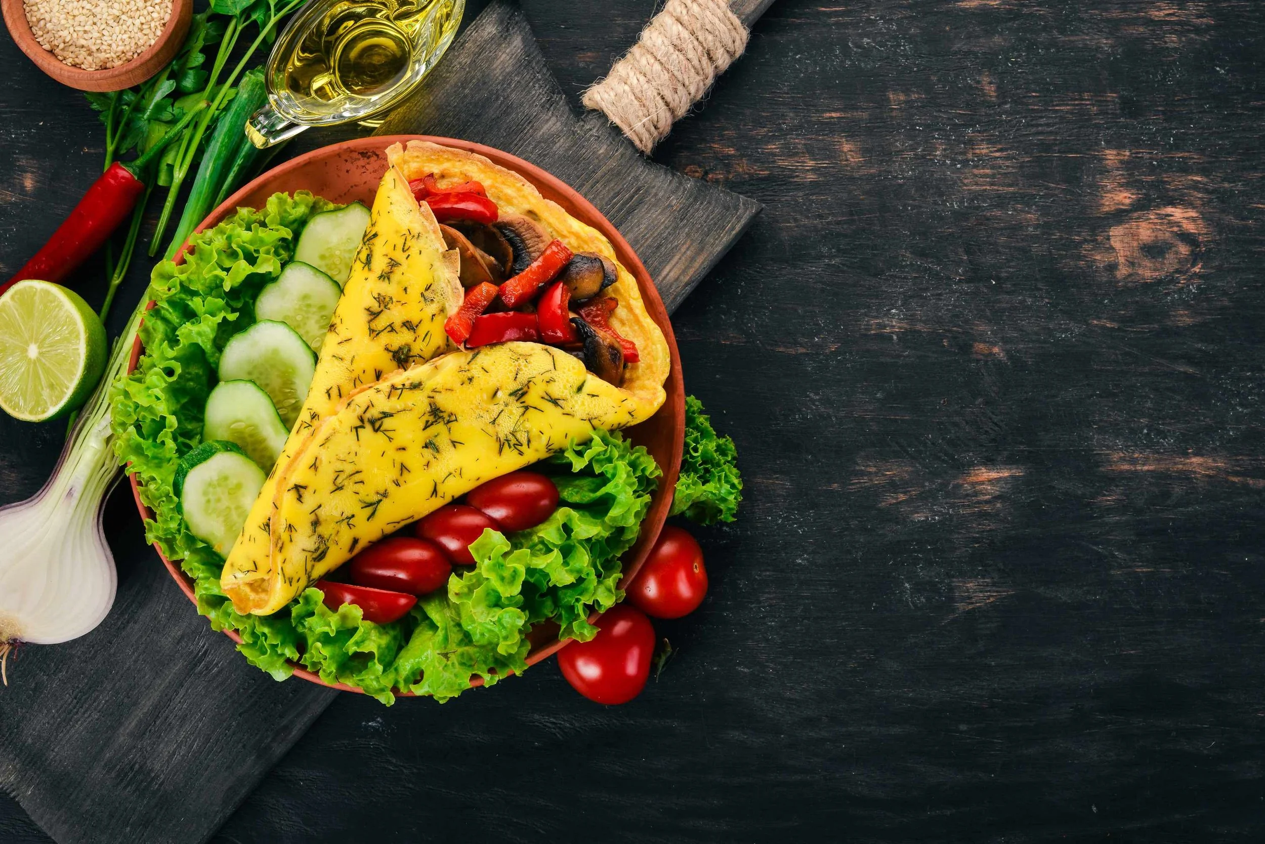 A bowl of colorful food including sliced cucumber, cherry tomatoes, a yellow omelet with herbs, grilled vegetables, and green lettuce, with a lime half, green onions, red chili, sesame seeds, olive oil, and a wooden spoon on a dark wooden surface.