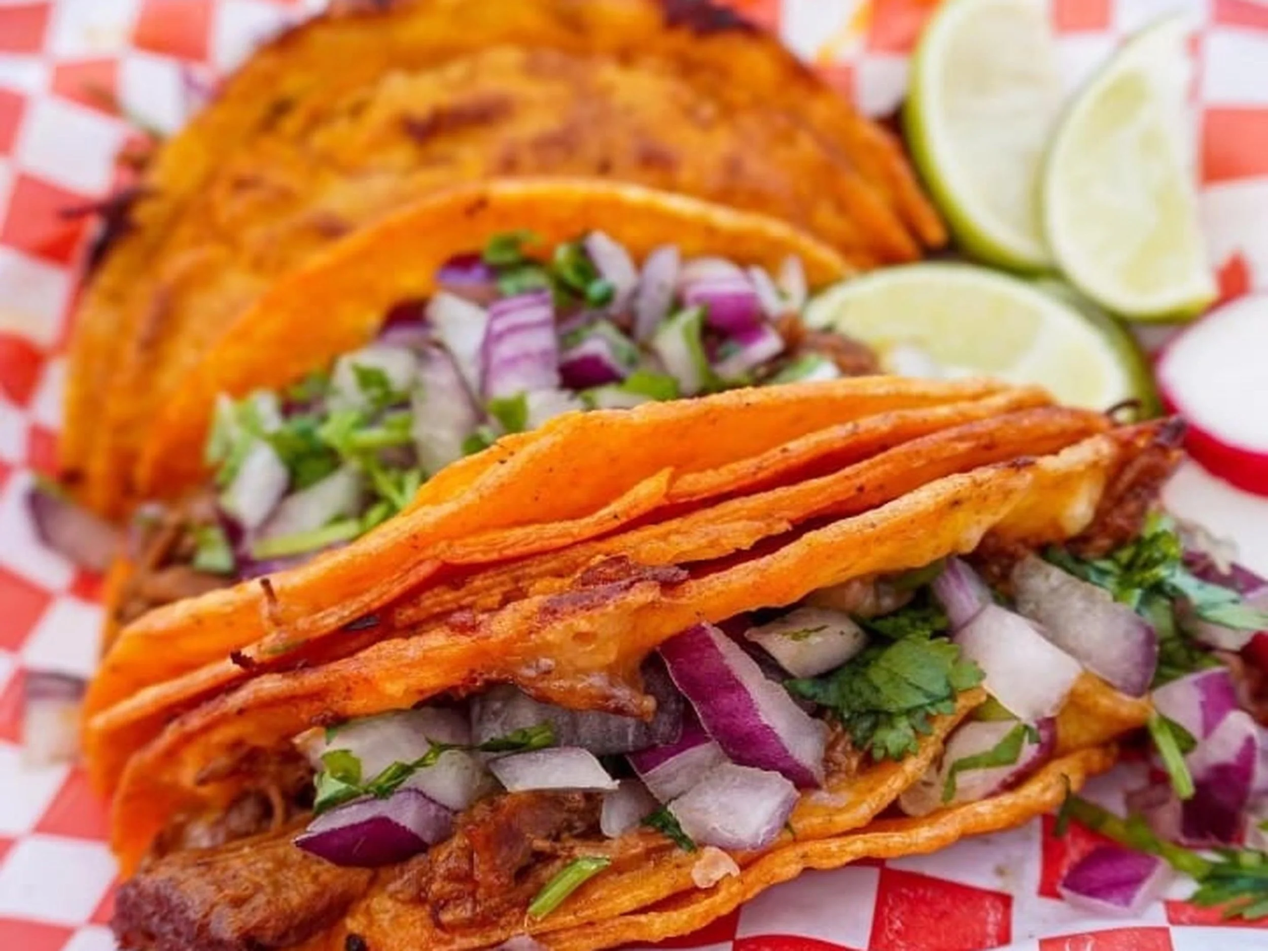 Close-up of a plate of tacos filled with shredded beef, diced onions, cilantro, and slices of lime, on checkered red and white paper.