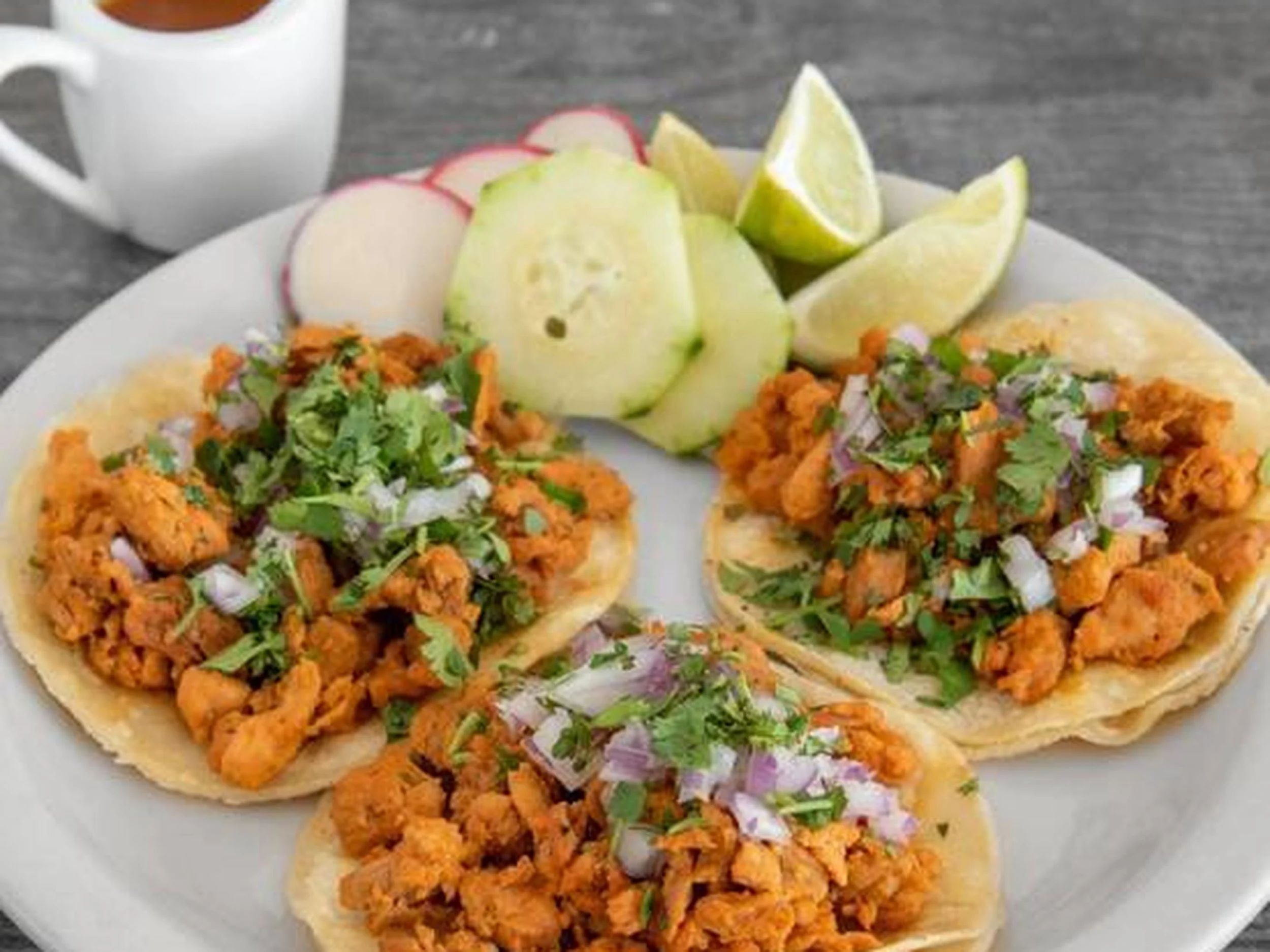 Plate with two tacos filled with seasoned meat, chopped onions, and cilantro, garnished with lime wedges and sliced radishes, on a gray wooden table with a cup of coffee in the background.