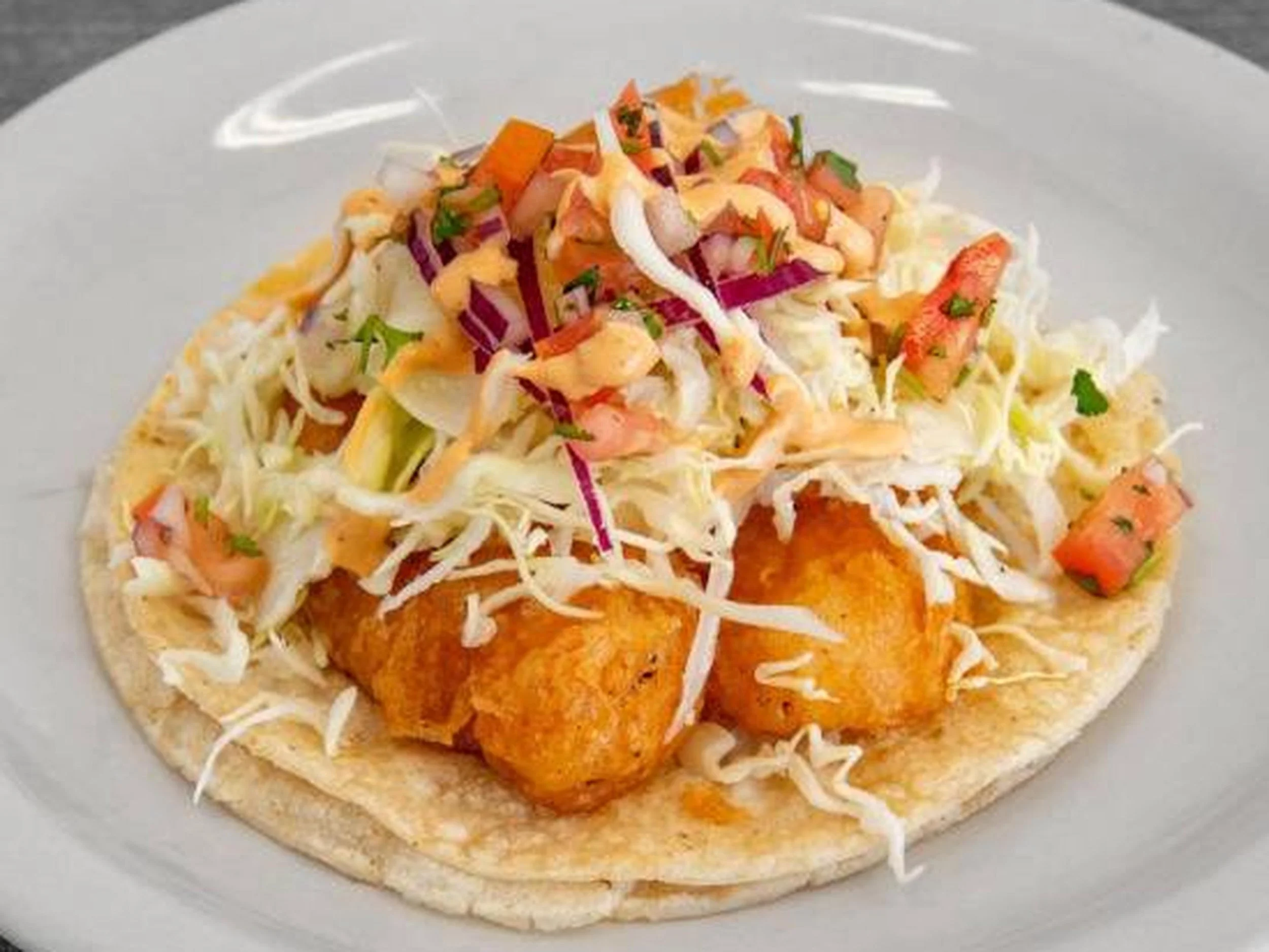A fish taco on a white plate topped with shredded cabbage, chopped tomatoes, chopped red onions, and fresh herbs.
