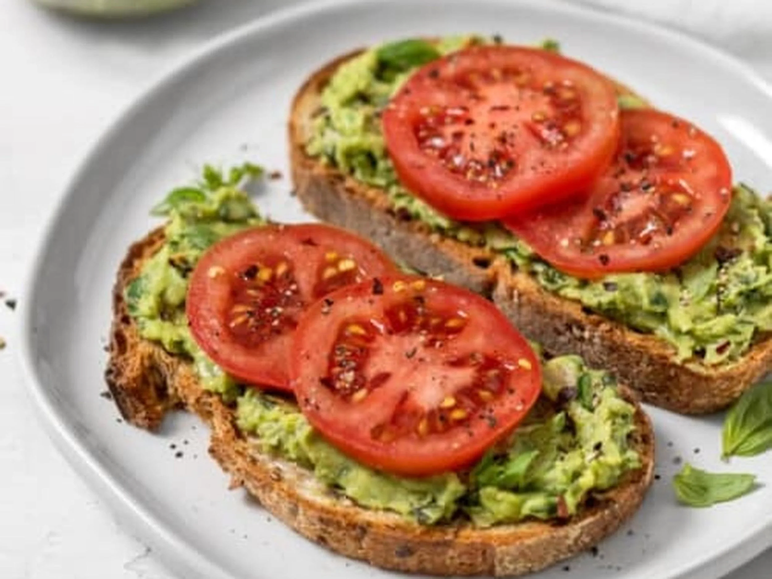 Slice of toasted bread topped with mashed avocado and sliced tomatoes, sprinkled with black pepper, on a white plate.
