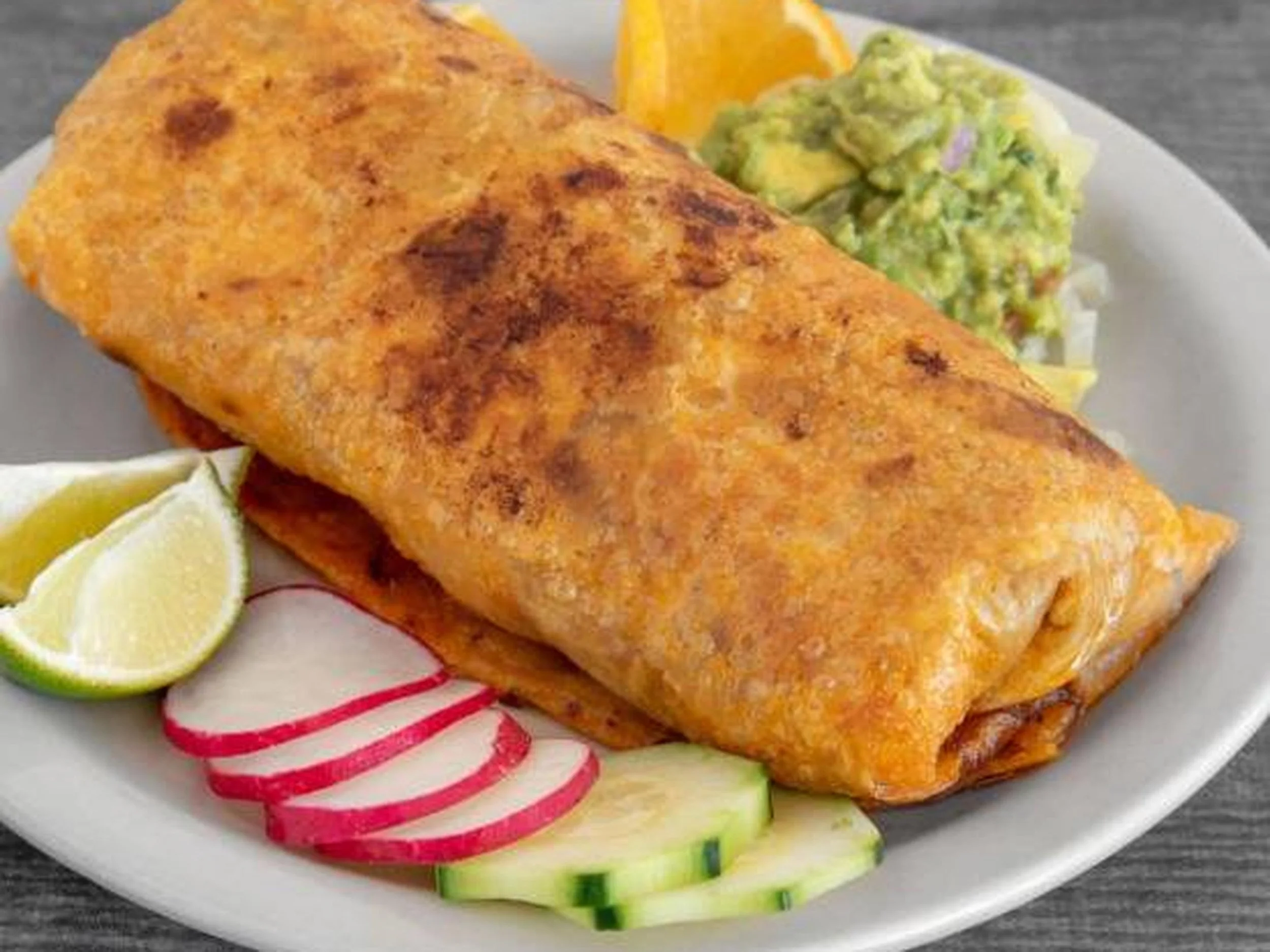 Fried fish fillet served with lemon wedges, sliced radishes, cucumbers, orange slices, and a side of avocado salad on a white plate.
