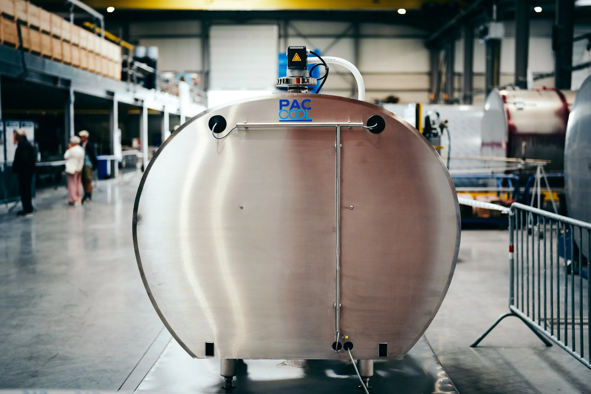 Large metallic tank in an industrial warehouse with a label 'PAC COOL' and a warning sensor on top, surrounded by barriers and people in the background.