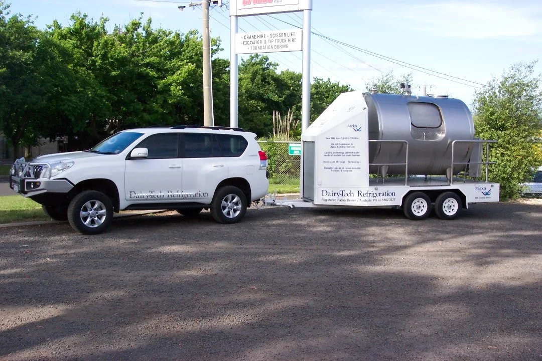 A white SUV towing a trailer with a large metal tank on it, parked on a gravel lot with trees in the background.