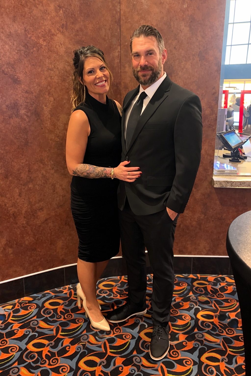 A smiling woman in a black sleeveless dress and beige high heels standing next to a man in a black suit, white shirt, and black tie, both posing indoors with a patterned carpet and a brown wall in the background.