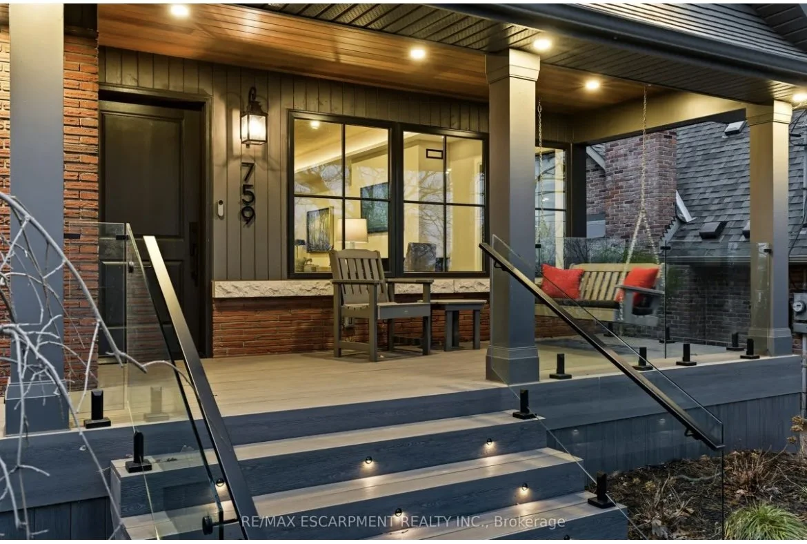 Front porch of a house with seating area, including a bench, chairs, and a swing with red cushions, illuminated by porch lights, brick and wooden exterior, glass railing, steps leading up to the porch.