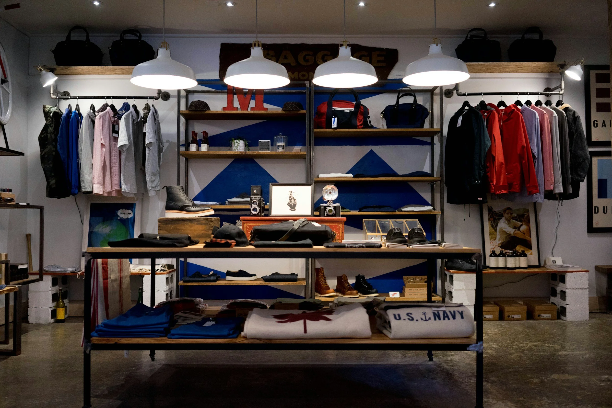 Clothing store display with shirts, jackets, hats, shoes, and bags arranged on metal and wooden shelves, illuminated by hanging white pendant lights.