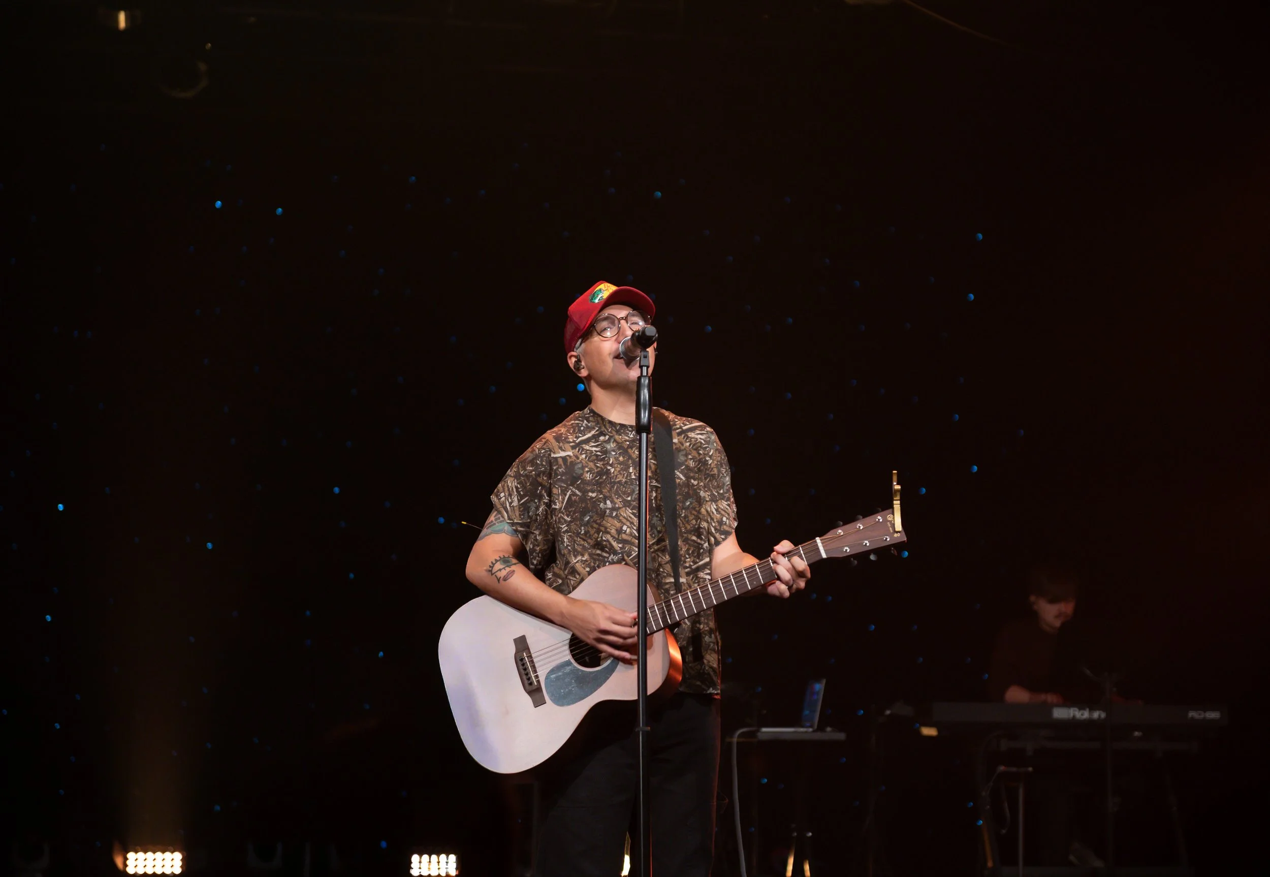 A young man wearing glasses, a red cap, and a camouflage shirt is singing into a microphone while playing an acoustic guitar on a stage with a starry background. There is a keyboard in the background.
