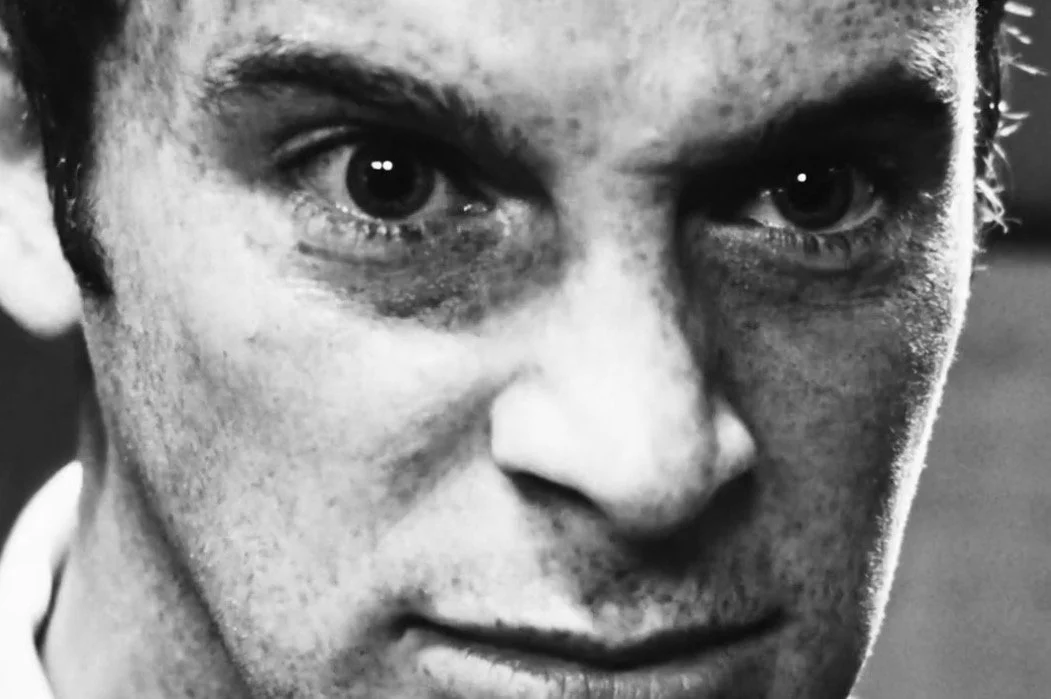 A close-up black and white photo of a man's face, showing intense eyes and serious expression.