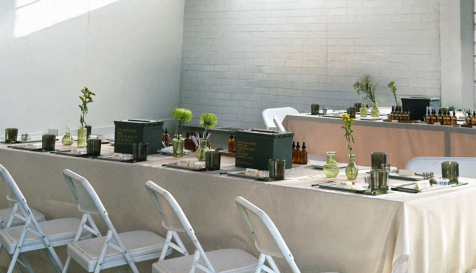 Long rectangular table set for a formal event with white chairs, decorated with small flower vases, candles, and bottles of essential oils or fragrances, against a white brick wall background.