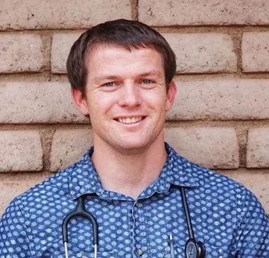 A smiling man wearing a blue patterned shirt and a stethoscope around his neck, standing in front of a brick wall.