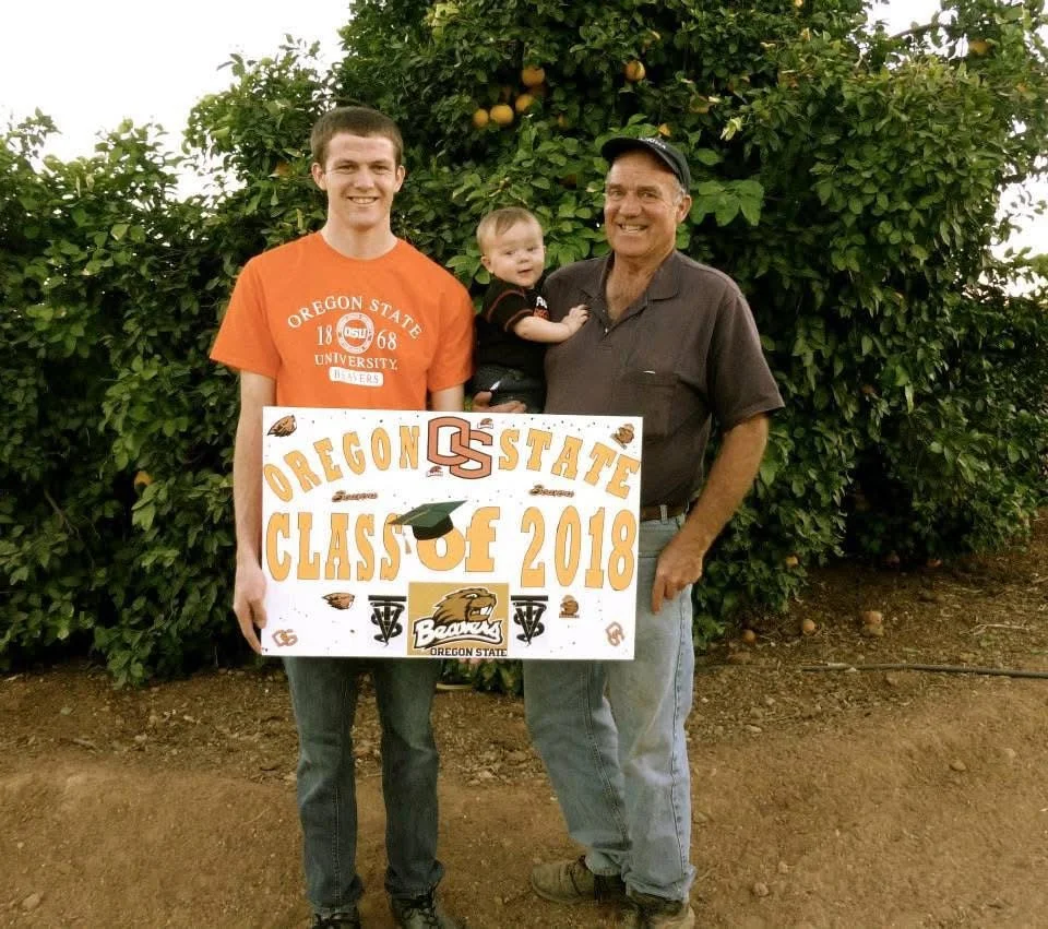 Three generations of a family celebrating a graduation in front of a tree with oranges, holding a sign that reads "Oregon State Class of 2018" with logos and a graduation cap graphic.