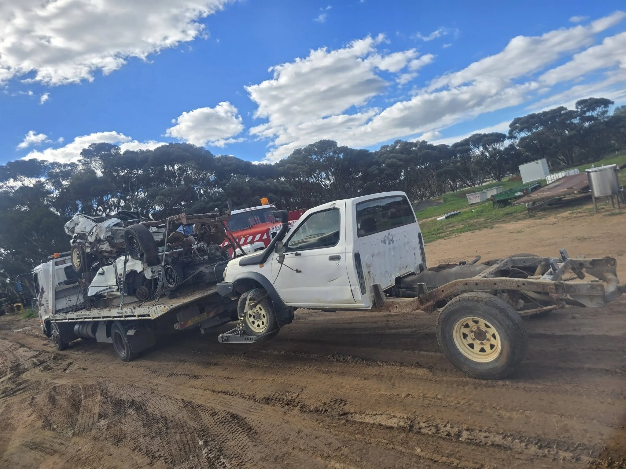 Image of several damaged vehicles wrecked on a lot with dirt ground and trees under a partly cloudy sky.