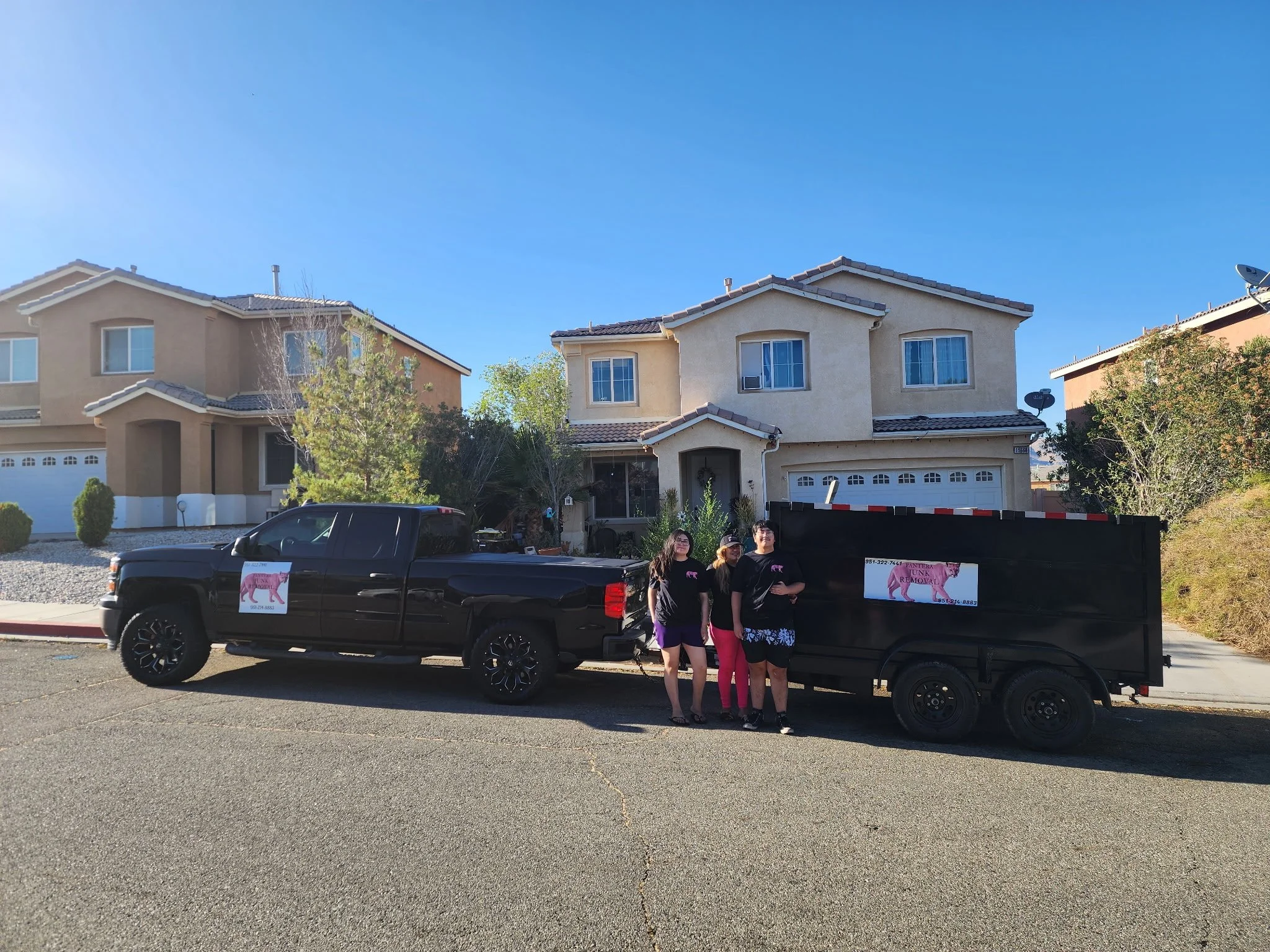 A group of four women standing in front of a black pickup truck with promotional signs on it, parked in front of two-story beige houses with tiled roofs and trees, under a clear blue sky.