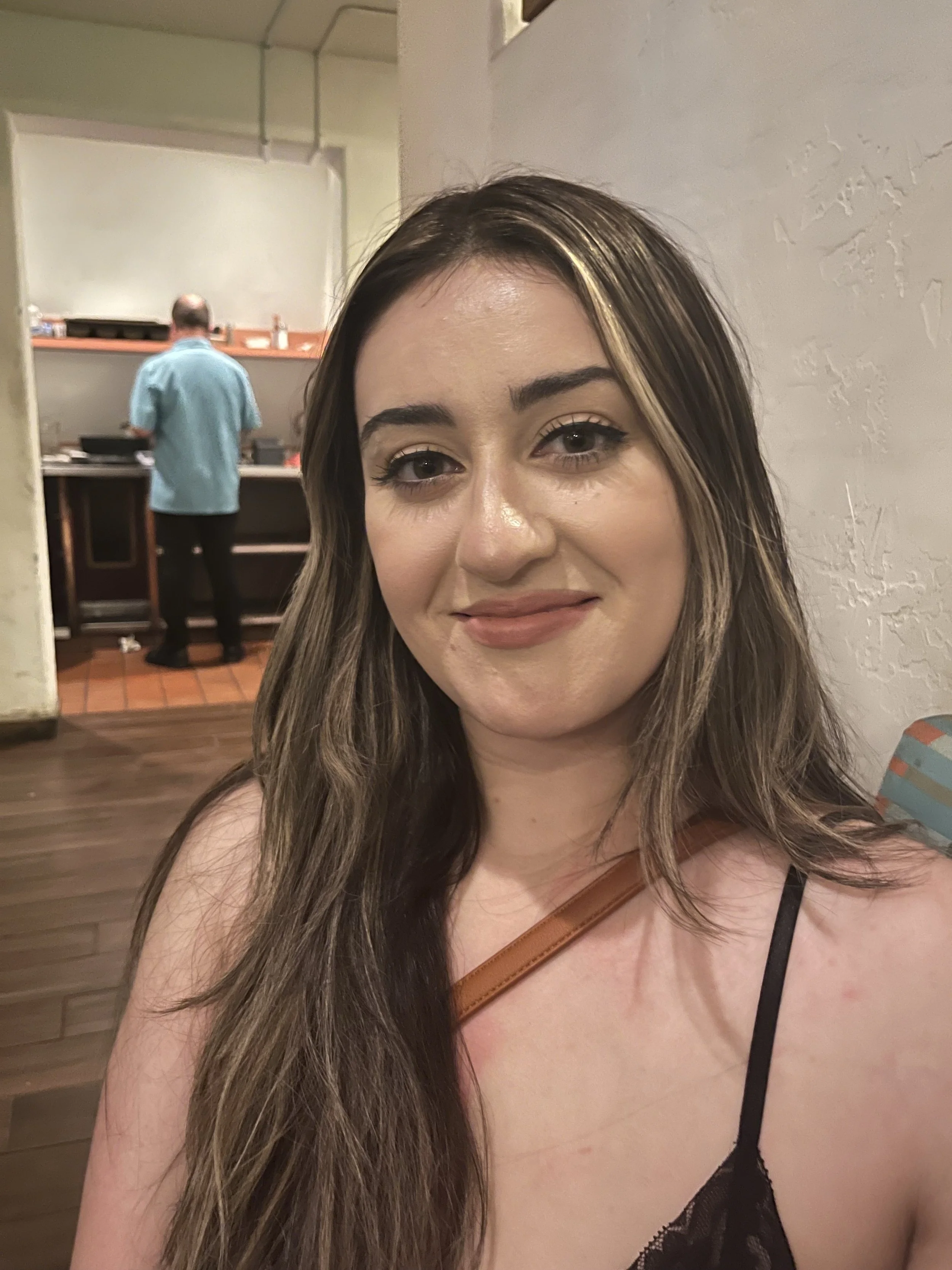 A woman with long brown hair and makeup taking a selfie in a restaurant or cafe, with a man in a blue shirt in the background near a counter.