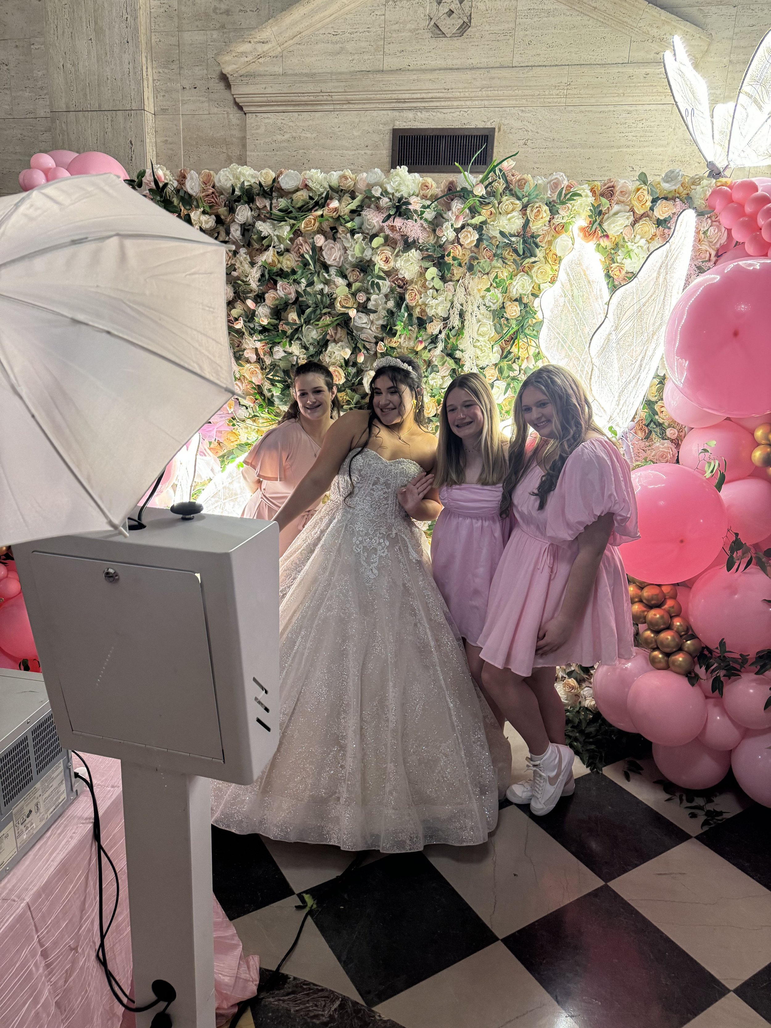 Group of four women, one in a wedding dress and three in pink dresses, standing together in front of a floral backdrop with pink and gold balloons, at a celebration or wedding event.