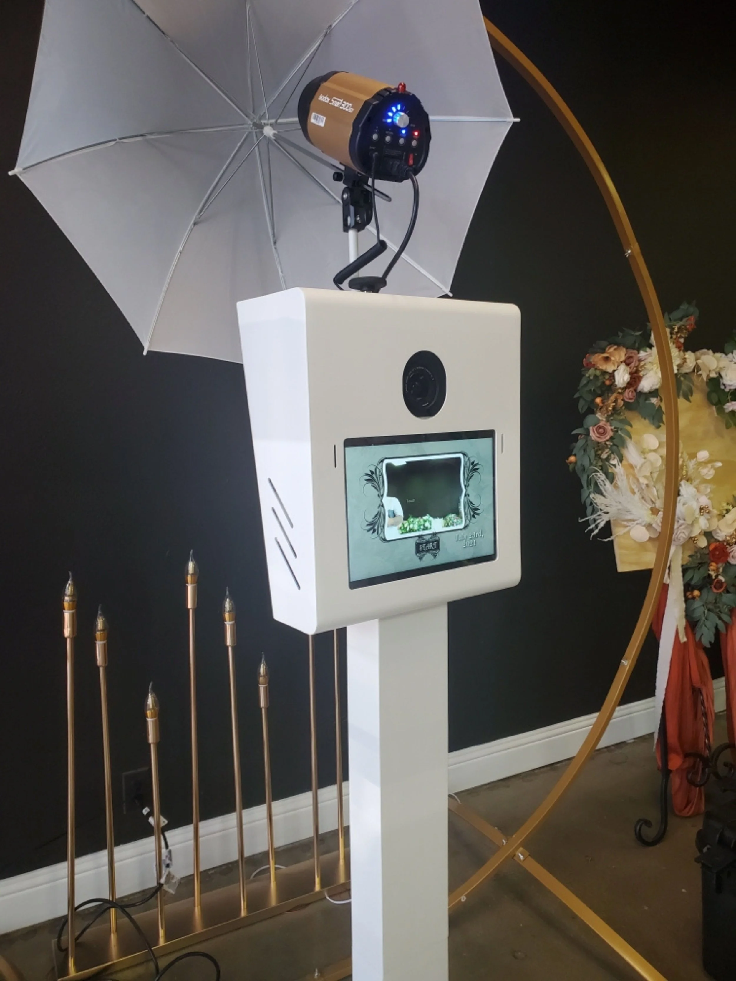 A photo showing a white stand with a small screen, a black camera, and a device with glowing blue lights mounted on an umbrella structure. Several gold-colored knife-like objects are aligned vertically in front of the stand, and part of a gold-colored arch and floral arrangement are visible to the right.