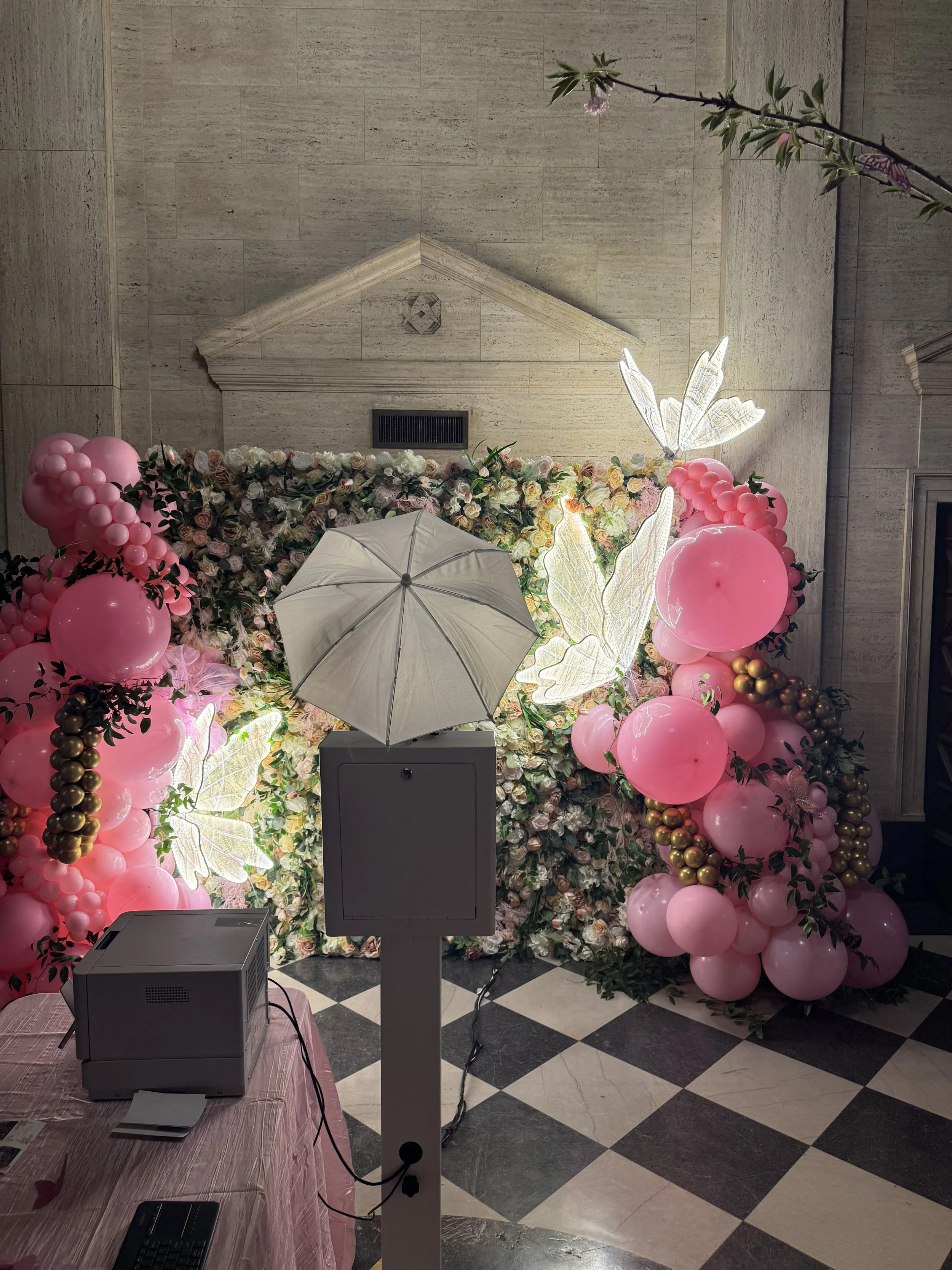 A decorated floral backdrop with pink and gold balloons, illuminated butterfly decorations, and greenery, set on a checkered floor with a gray air vent on a beige wall in the background.