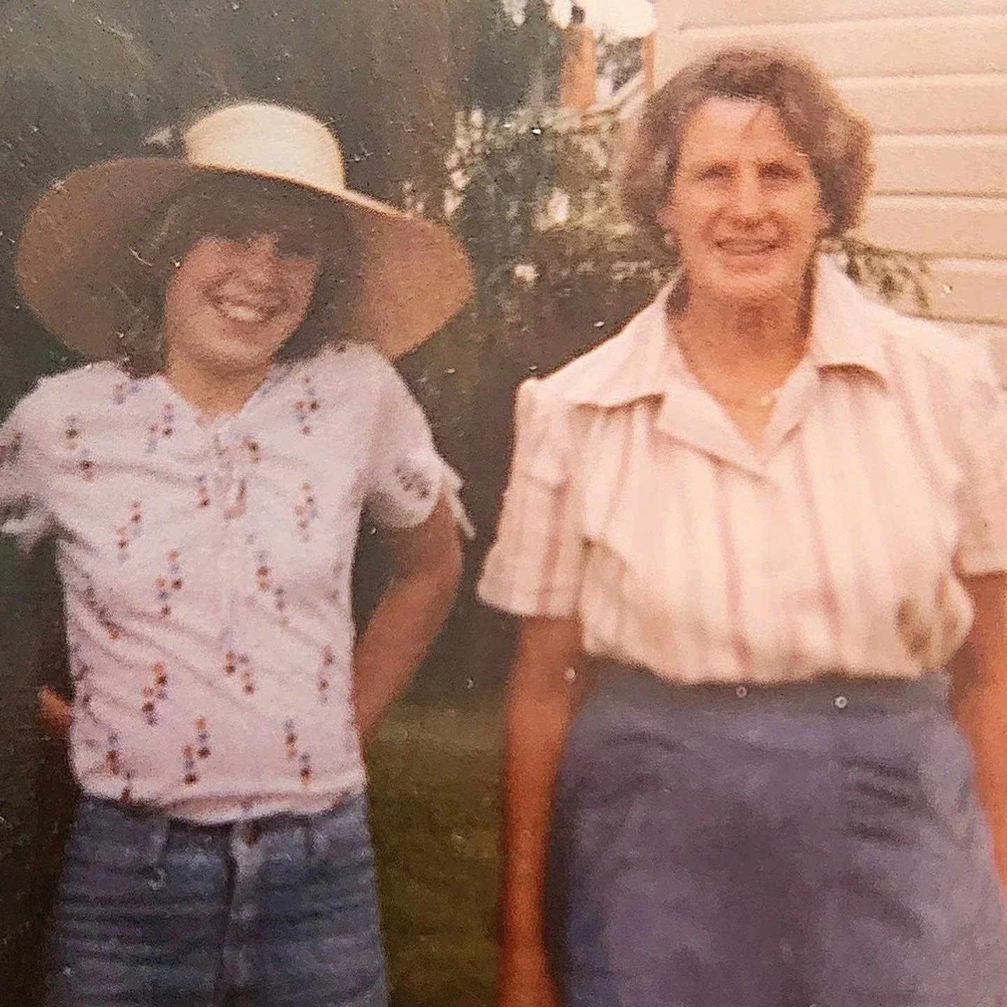 A young girl wearing a white floral blouse, blue jeans, and a large straw hat, and an older woman wearing a striped short-sleeve shirt and a dark skirt, smiling outdoors with a house and trees in the background.