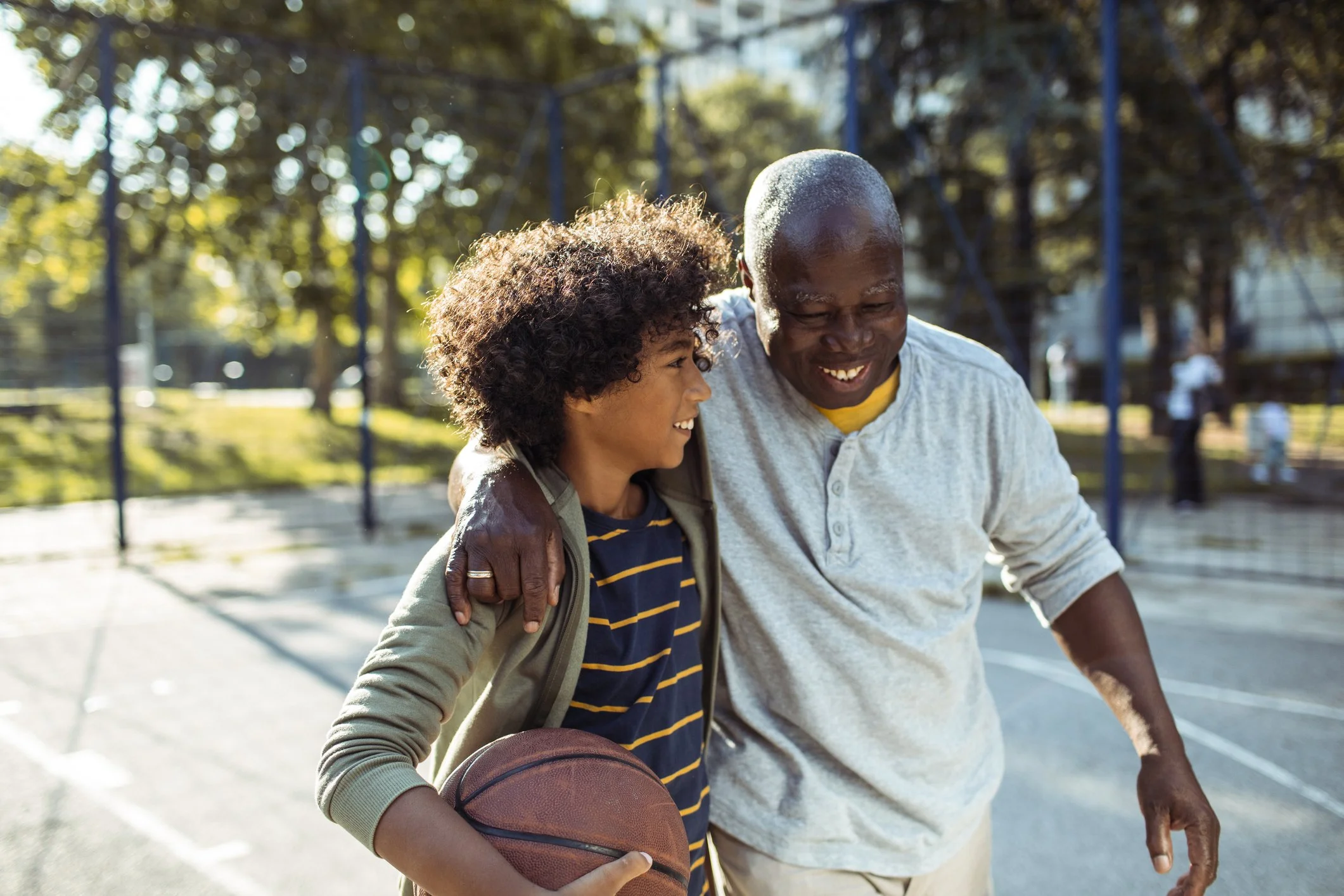 A happy African American man and woman sharing a moment at the basketball court, with the woman holding a basketball. The man has his arm around the woman's shoulder and they are smiling at each other. The background shows trees and a blue fence.