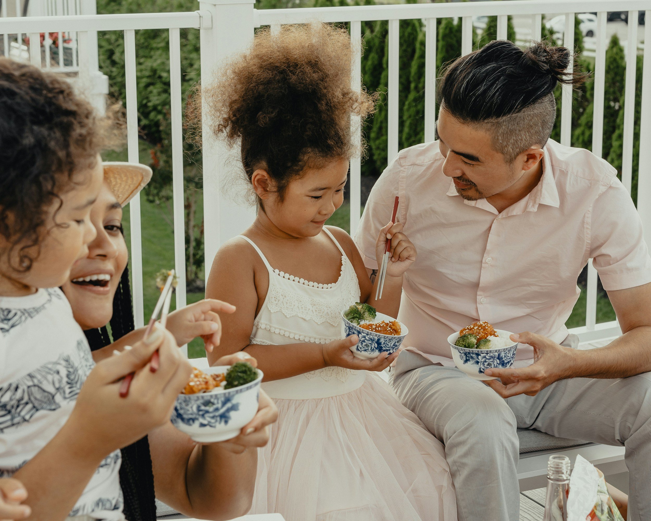 A group of people, including children and an adult male, seated outdoors near a white fence, eating from bowls of food, with greenery in the background.