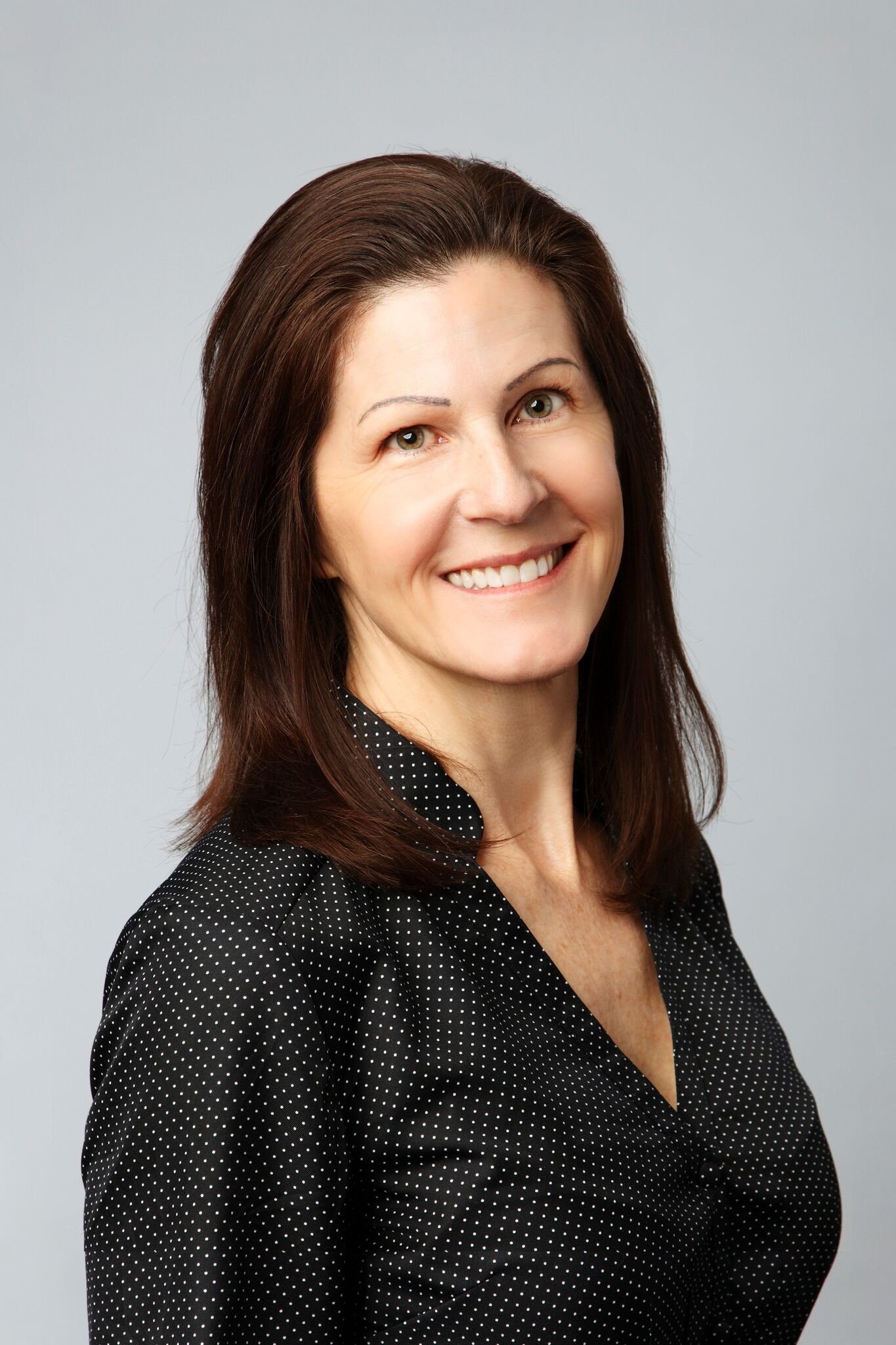 Portrait of a smiling woman with brown hair, wearing a black polka-dotted blazer, against a plain light gray background.