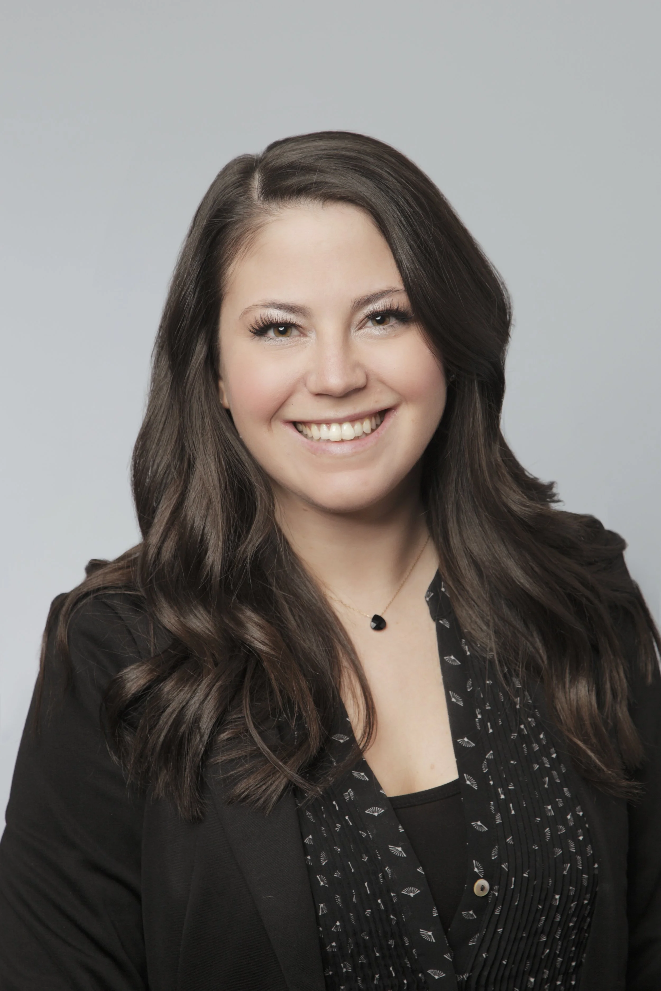 A woman with long dark hair smiling, dressed in a black blazer and patterned top, against a light gray background.