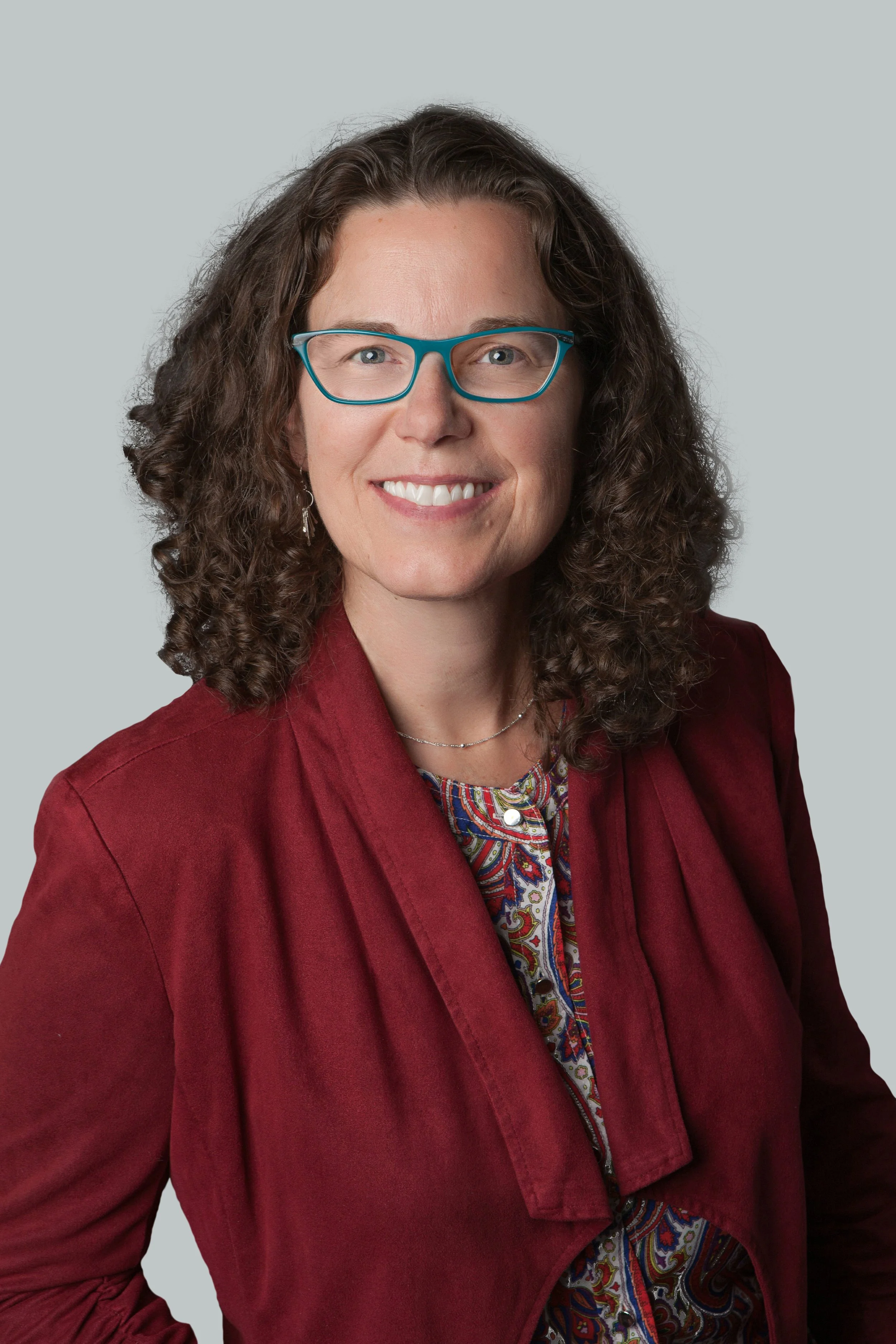 A woman with curly brown hair, wearing teal glasses, a red blazer, and a colorful patterned shirt, smiling at the camera.