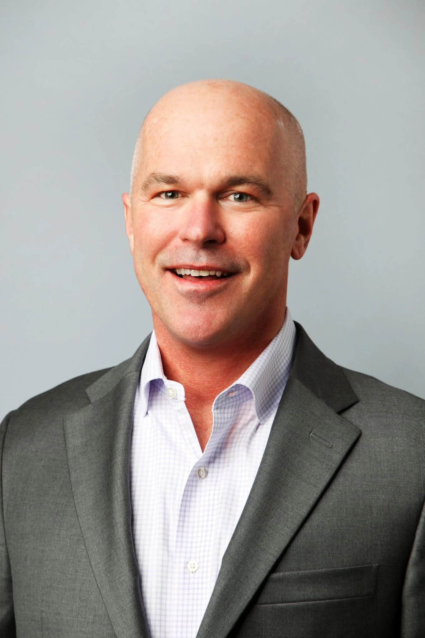 A man with a bald head wearing a gray suit and white shirt, smiling against a plain, light gray background.