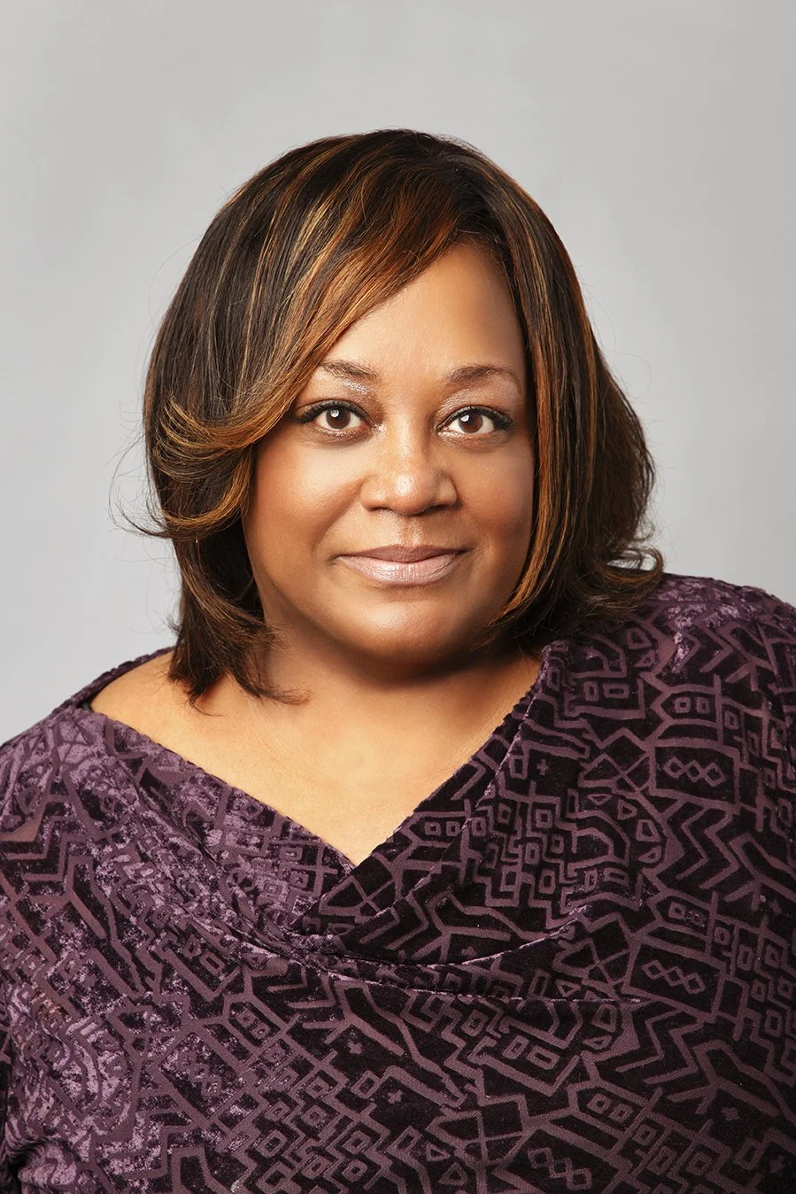 Headshot of an African American woman with shoulder-length hair, wearing a purple patterned top, against a light grey background.