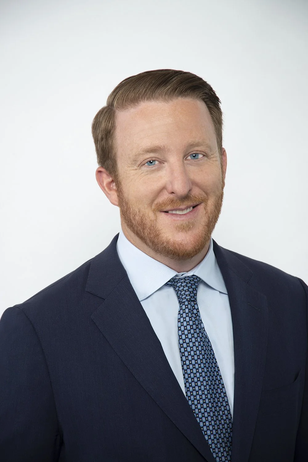 Professional headshot of a smiling man with red hair and beard, wearing a dark suit, light blue shirt, and patterned tie against a plain background.