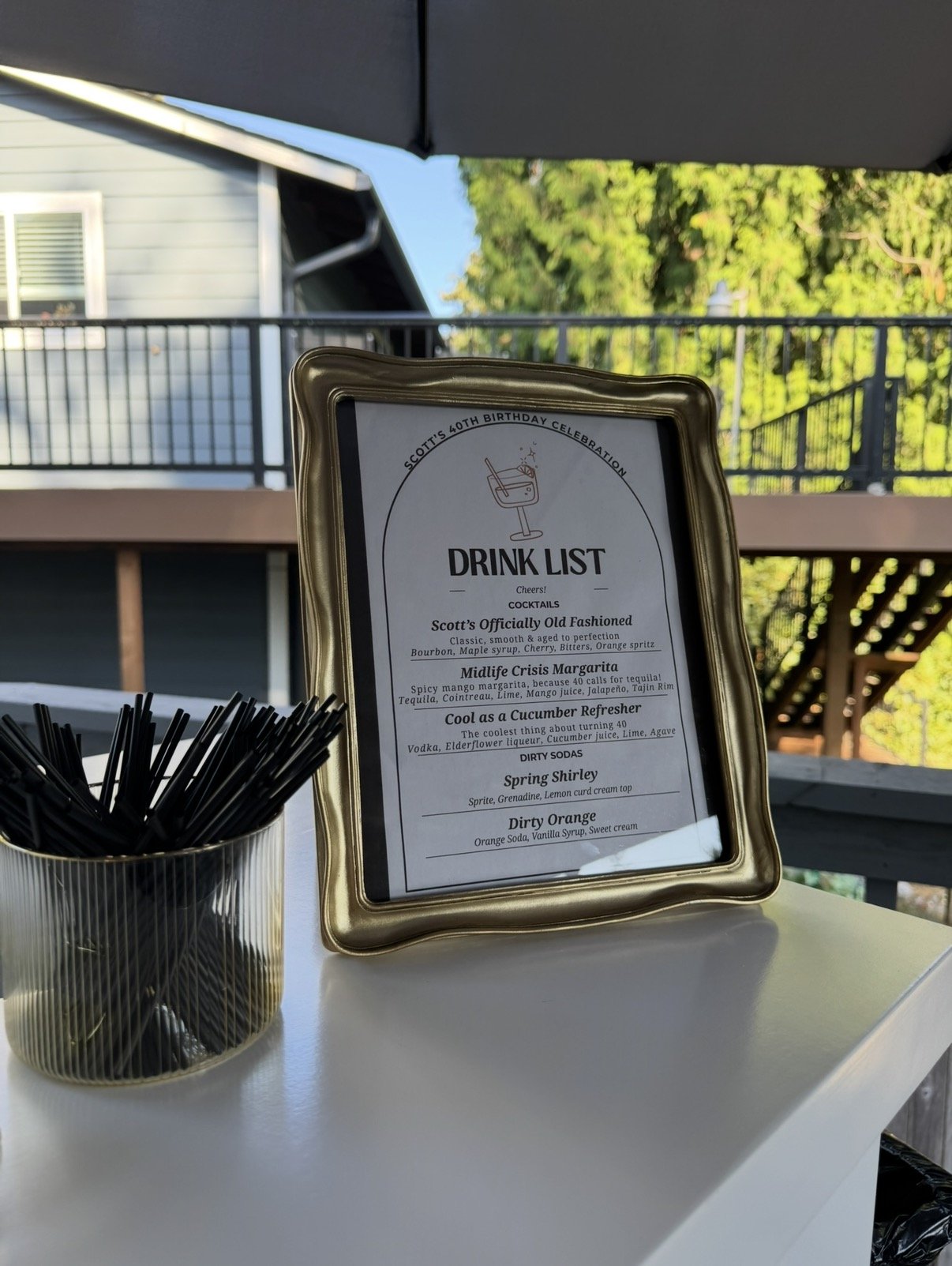 A drink menu on a mobile bar setup for a birthday party with a container of black straws, patio in the background, a house, and green trees in the background.