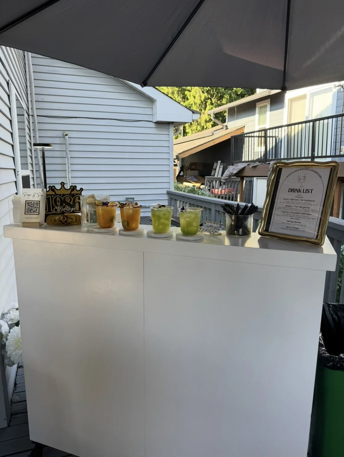 Outside drink display table with various colorful cocktails, drink list frame, umbrella, and background of houses and trees.