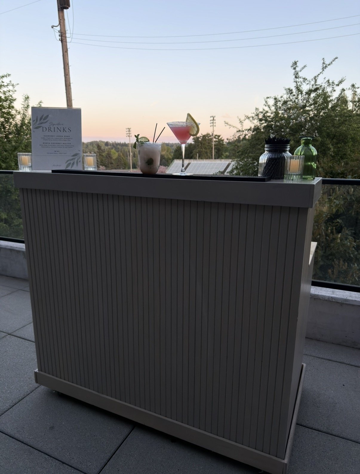 Outdoor bar with drinks on top, including a cocktail with lemon wedge, a drink with straw, candles, and greenery in the background at sunset.