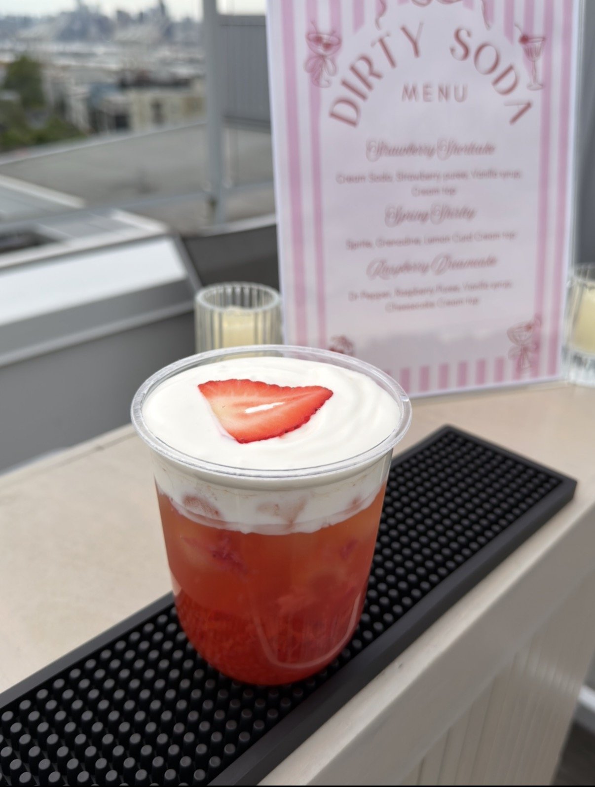 A strawberry cream soda with cold foam and a strawberry slice on top, placed on a bar mat on a ledge. In the background, there is a pink and white striped menu sign with desserts and drinks listed, and a cityscape in the distance.