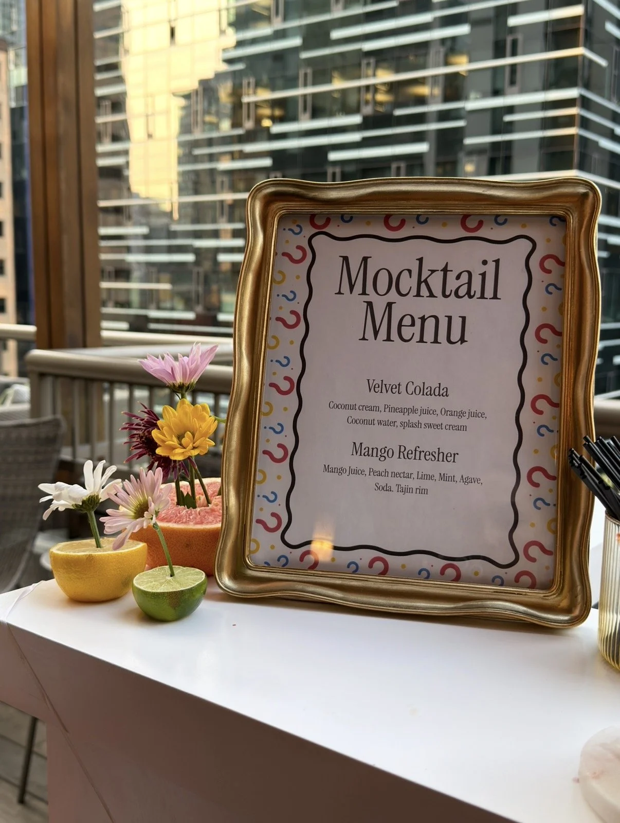 A framed mocktail menu featuring two drinks, Velvet Colada and Mango Refresher, on a white table with colorful floral decorations including a lemon, lime, and a watermelon half adorned with flowers, with an urban cityscape in the background.