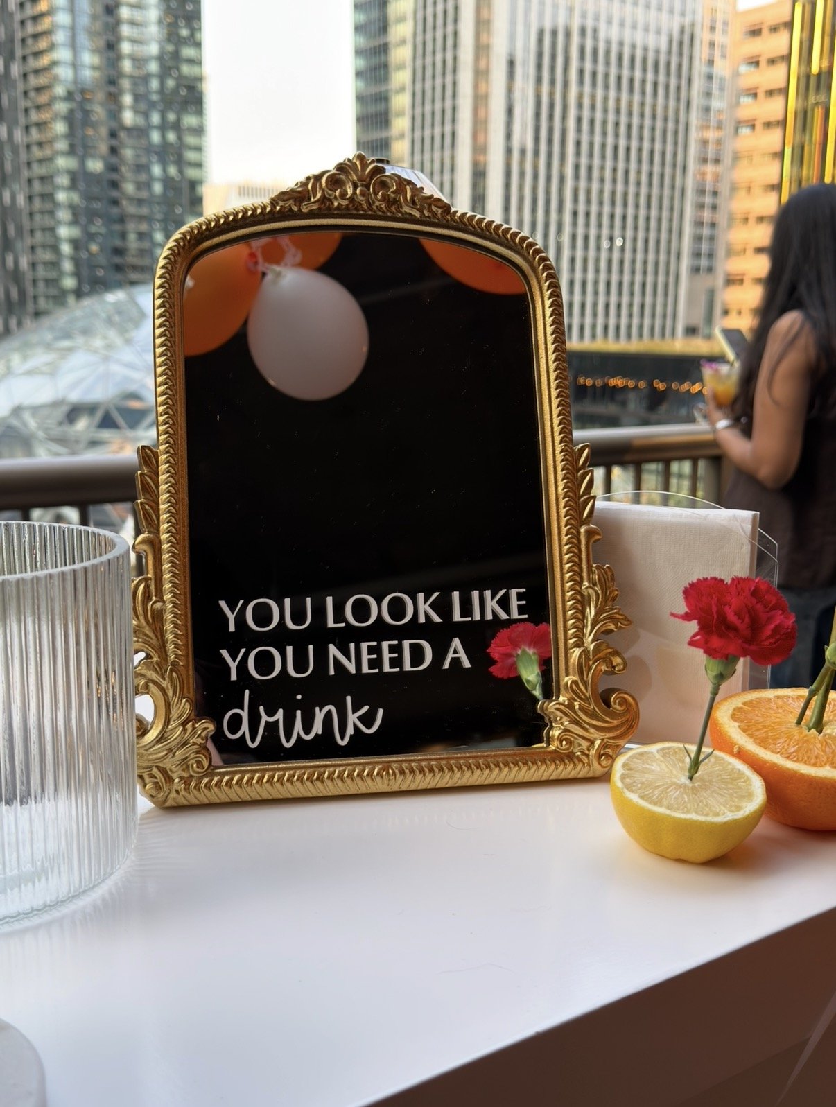 A decorative mirror with a message that says, 'You look like you need a drink,' at an outdoor party or gathering, with a city skyline in the background, a glass of orange drink with a straw, and half an orange and lemon with mini flowers as table dec