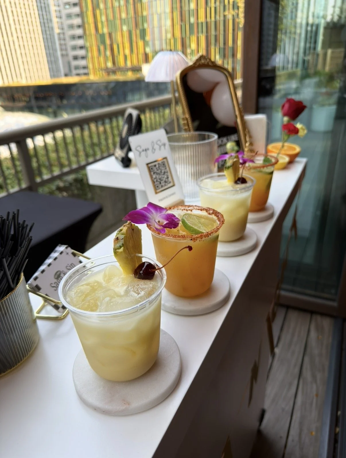 Four colorful cocktails garnished with fruit slices and flowers, arranged on a white table on a balcony with a cityscape view of Seattle, WA.