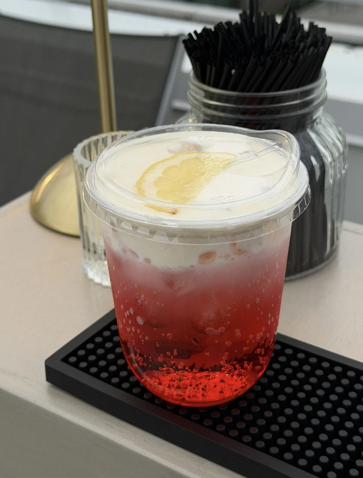 A sprite based dirty soda with a lemon curd cold foam and lemon garnish, placed on a black tray, with a jar of black straws and a glass of water in the background.