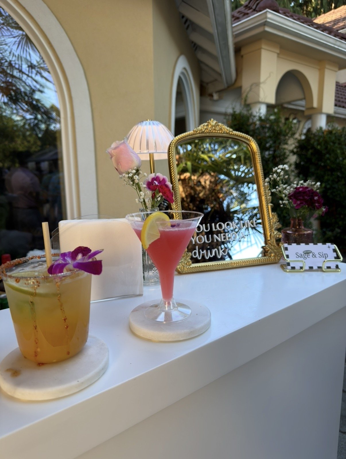 A display of three cocktails with floral garnishes, a mirror with the text "You look like you need a drink," and decorative flowers on a white table outdoors.