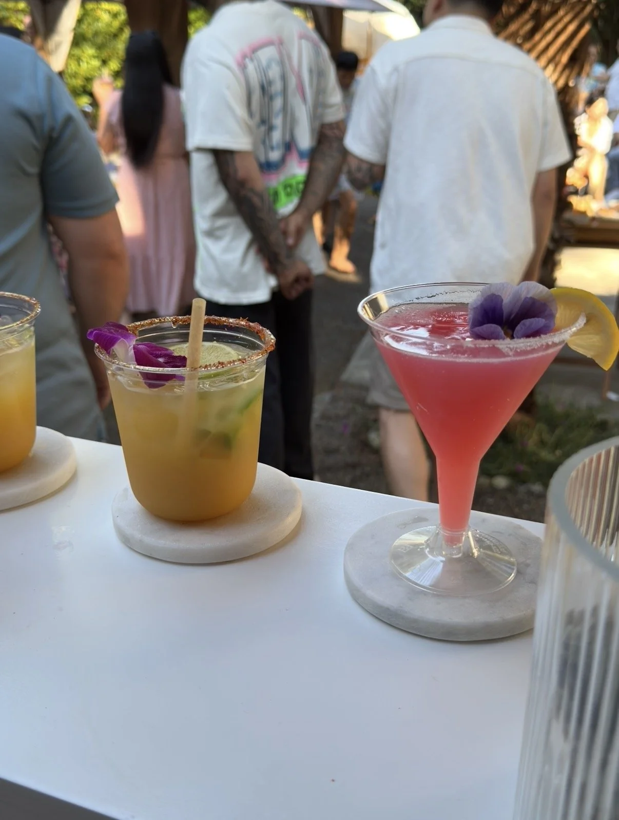 Colorful cocktails on a white table with blurred people in the background at an outdoor gathering.
