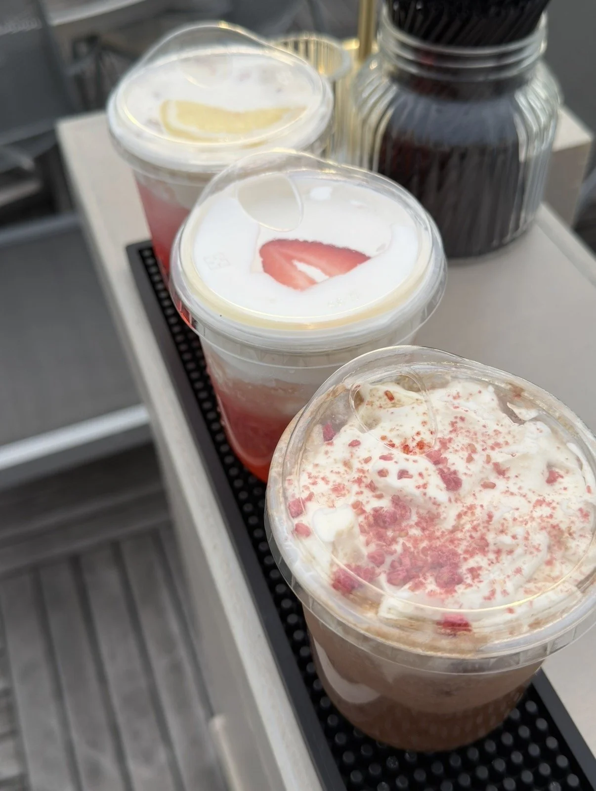 Three cold beverages in plastic cups with domed lids, topped with whipped cream and pink sprinkles, on a black tray. Two small jars containing dark liquids are in the background.