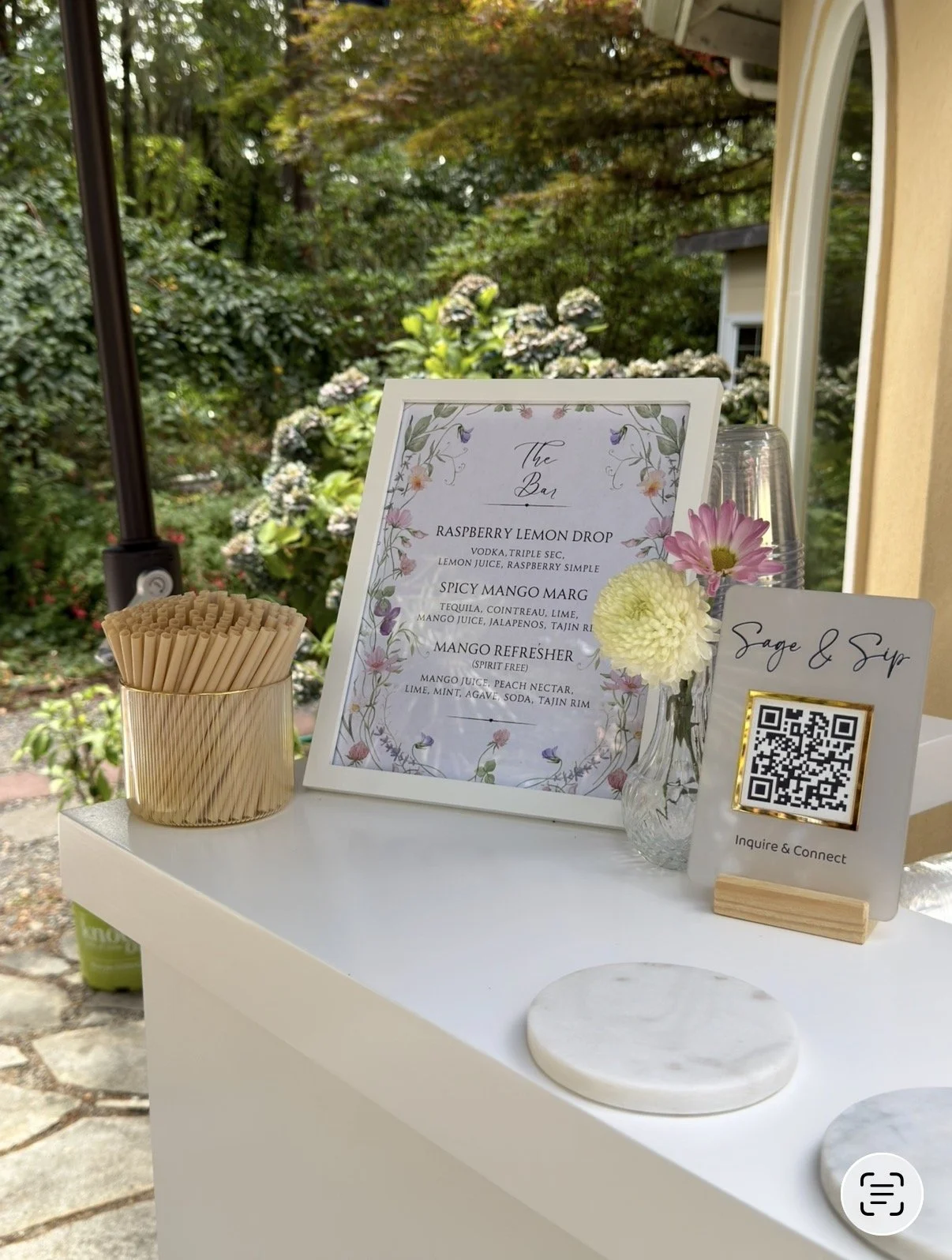 A table with a floral menu, a flower vase, a container of wooden stir sticks, and a QR code sign for Sage & Sip in an outdoor garden setting.