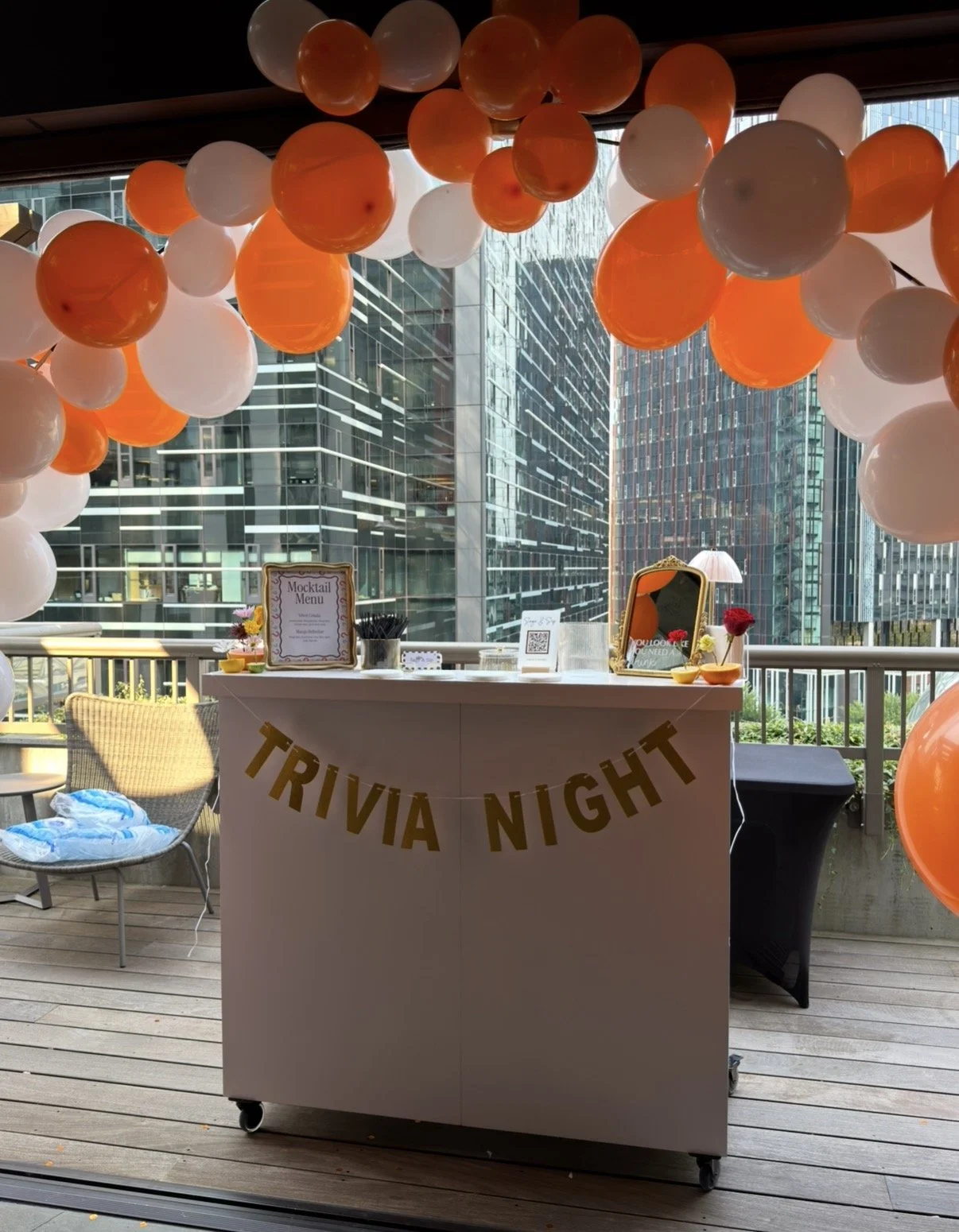 A decorated space with orange and white balloons forming an arch, a table with a "Trivia Night" banner, and a city view through large windows in the background.
