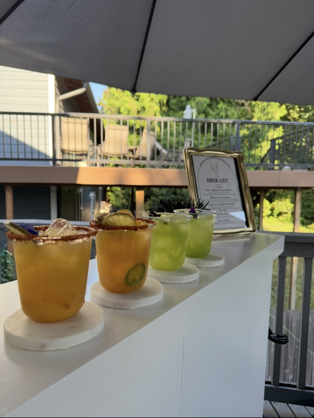 Four colorful cocktails with garnishes lined up on a white bar counter, with a drink list menu and a balcony with outdoor furniture in the background.